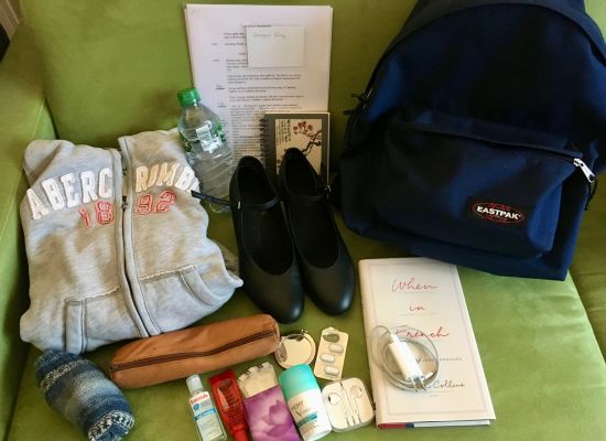 Items for a rehearsal bag laid out on a green couch: grey hoodie, woollen socks, pencil case, lip balm, deodorant, headphones, charger, sheet music, book (When in French by Laura Collins), black character shoes, notebook, drink bottle, tissues, plasters, hand sanitiser