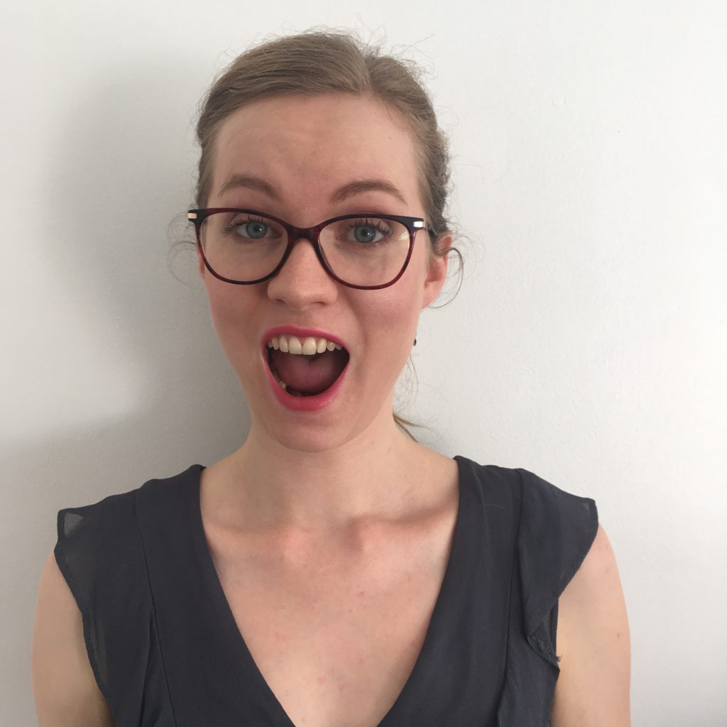 Georgia Aussenac is stretching her mouth wide open, and pulling a silly face. She's wearing bright pink lipstick and red framed glasses, and has her hair tied back.