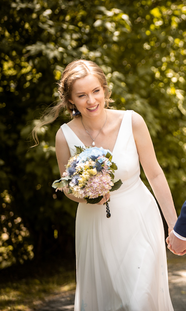 Georgia is wearing a white wedding dress, holding a blush pink and pastel blue bouquet, against a backdrop of summer light through tree leaves. She's smiling with pure joy.