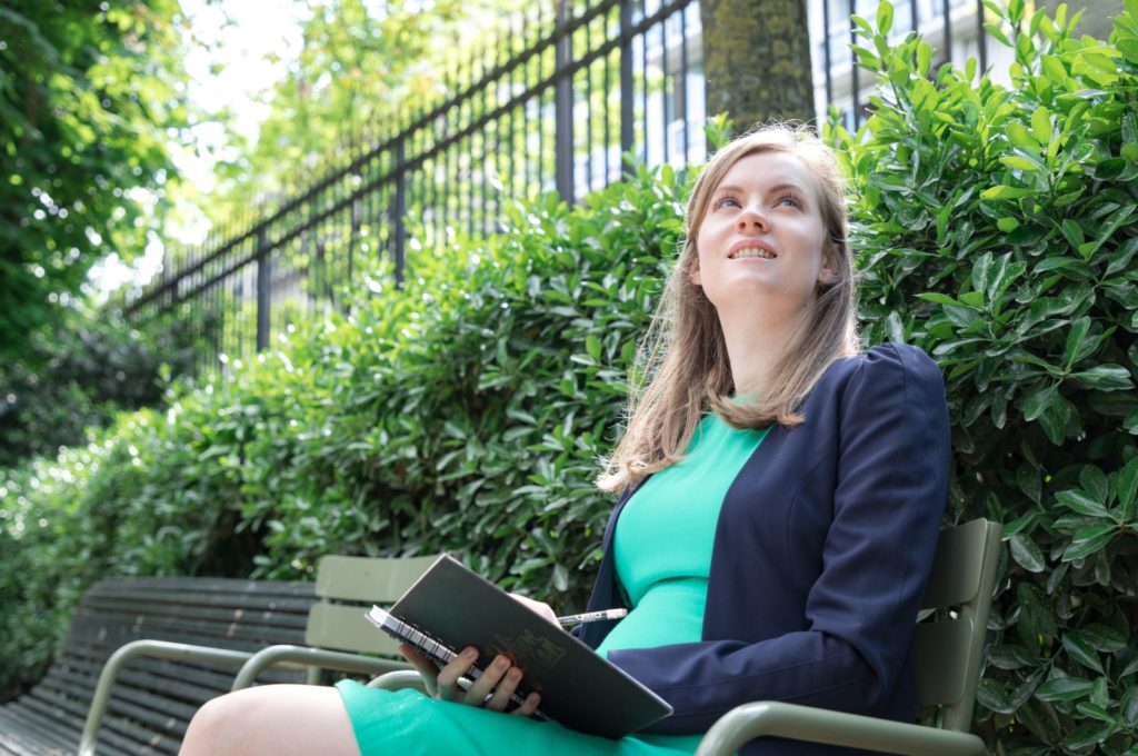 Georgia, a young blonde white woman is sitting on a bench in a park with a journal in her hand looking inquisitively towards the sky. She is likely reflecting on how best to trust your gut when it comes to singing.