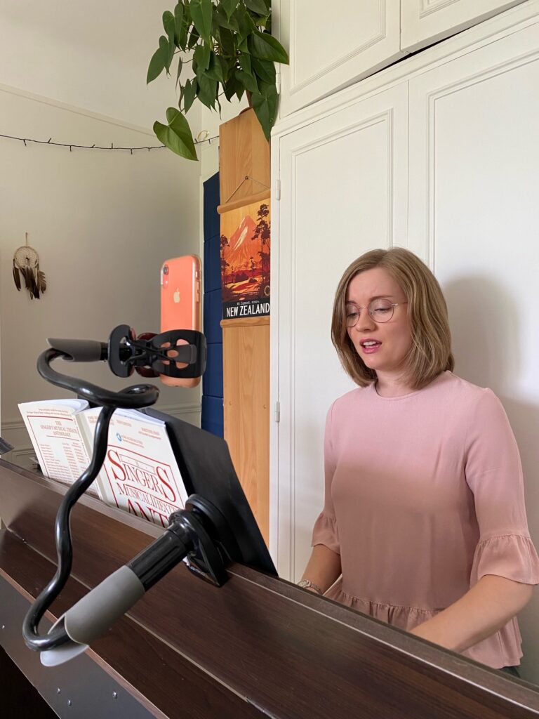 Georgia, a white woman in her late 20s is playing the piano and singing. She has shoulder length blonde hair, pink wire rimmed glasses and is wearing a light pink top. She has just completed her warm up for singing and she is ready to begin her singing practice