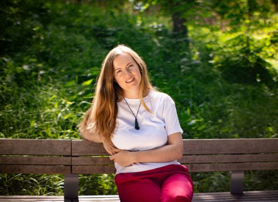 Georgia, a white woman in her 30s with long blonde hair is sitting on a bench in a park, leaning on the back rest with one arm and smiling directly at the camera.