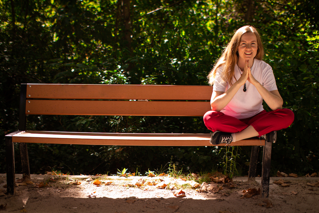 Georgia is sitting cross-legged on the end of a bench in a park, resting her chin on her hands and smiling at the camera