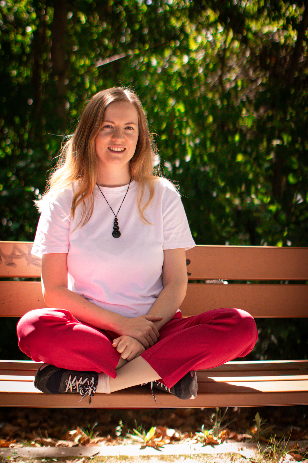Georgia, a white woman with blonde hair is sitting cross-legged relaxed on a park bench, smiling at the camera. She is wearing a white tshirt, her pounamu necklace, and hot pink trousers. GL Voice Singing Coach