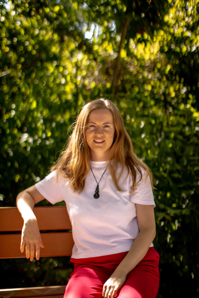 Georgia, a white woman with long blonde hair in her 30s is sitting on a bench, leaning on one arm and looking directly at the camera. She's wearing a white tshirt and her pounamu necklace.