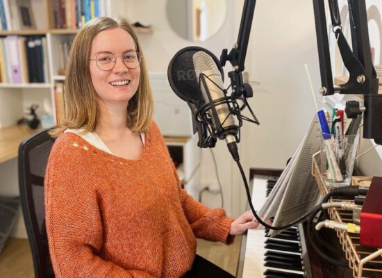 Georgia is sitting with one hand on her keyboard, in front of her microphone. She's wearing a fluffy orange jumper and black leggings, and is smiling.
