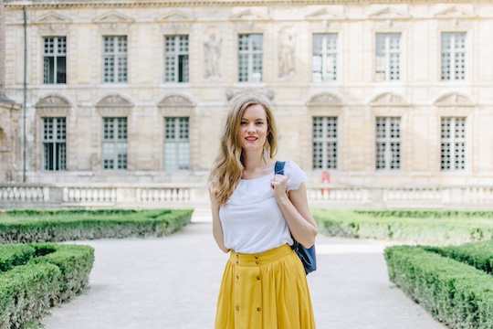 Georgia is standing in a renaissance French courtyard, wearing a white lacy top and a high waisted mustard yellow circle skirt. She's got on a navy blue backpack, and she's holding on to one of the straps.