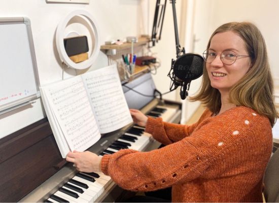 Georgia Aussenac sitting at her piano, holding open a book of sheet music, ready to coach. She's wearing a big fluffy orange mohair jersey, and she's smiling.