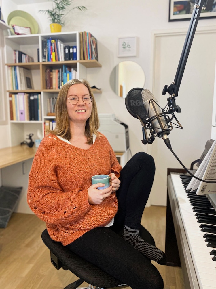 Georgia Aussenac is sitting at her piano in her home studio, wearing a fluffy oranger jumper and black leggings, and holding a mug of something hot.