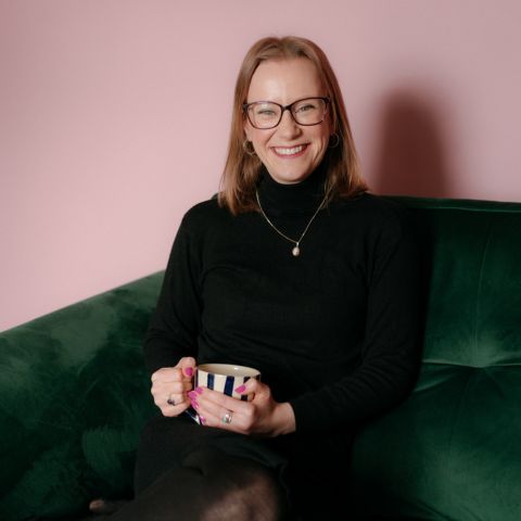 Cara Redpath is sitting on a green velvet couch, with a blush pink wall behind her. She's wearing all black, which contrasts well with her dark blonde hair and dark framed glasses. She's got an exciting smile, and she's holding a cup of tea.