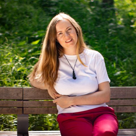 Georgia Aussenac is sitting on a bench in a park, leaning back on one elbow. She's wearing a white tee and hot pink trousers, and her pounamu necklace. Her long blond hair is loose, and she's smiling.