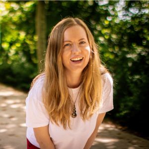 Georgia Aussenac, a white woman with long blonde hair, is leaning forward into the camera, hands on knees, laughing. She's wearing a white tee, and standing on a path outdoors.