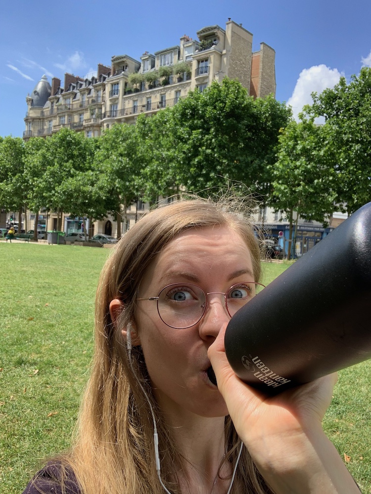 Georgia Aussenac sitting in a parisian park, drinking from a black sports drink bottle. She's got headphones in, is wearing glasses, and is pulling a 'have you drunk enough water today' face. There is green grass, blue skies, plane trees and typical Parisian building in the background.