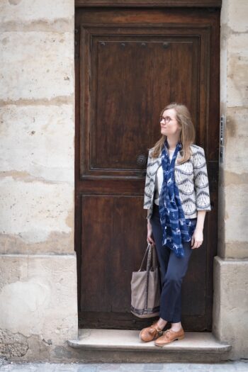 Georgia Aussenac is standing in front of a old wooden door, outside a stone parisian building. She's carring a tote bag with silver sequins, and wearing a cute geometric jacket, blue slacks and brown leather plimsoles. She looks young and curious.