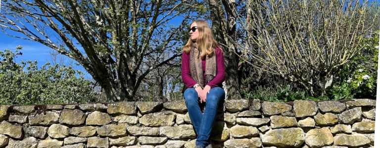 Georgia Aussenac is sitting on an old stone wall, looking at the view. She is wearing a dark pink knit sweater, blue jeans and sun glasses, and is surrounded by a garden of plants blossoming and budding for spring.