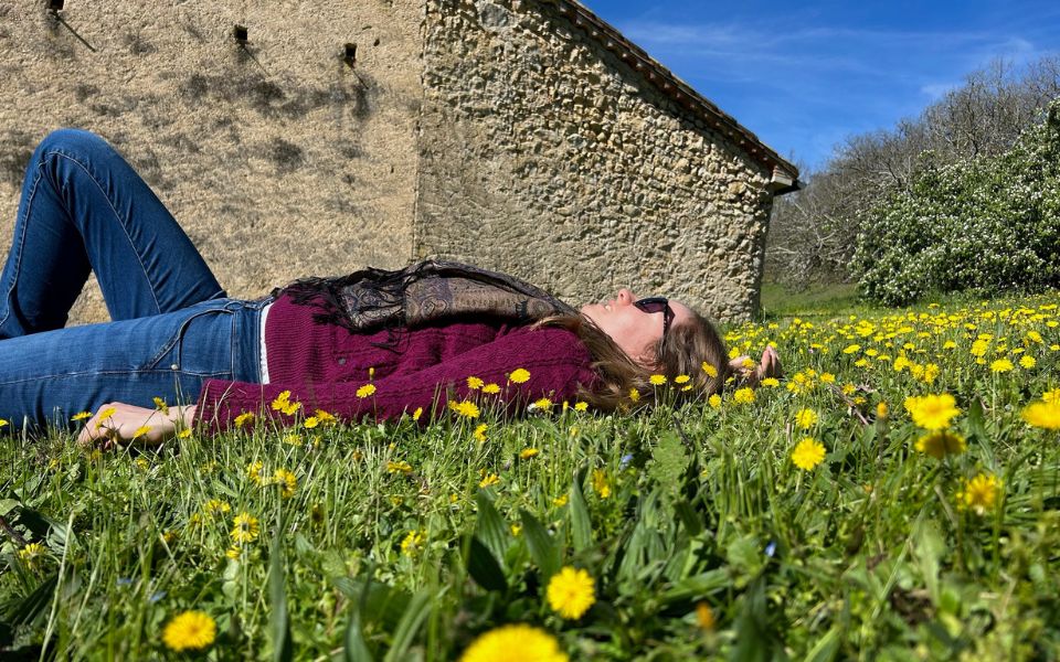Georgia Aussenac is lying on a lawn surrounded by yellow daisies, in front of an old stone farm building. She's wearing a dark pink knit sweater, jeans and sunglasses, and she's relaxing looking up at the blue sky.