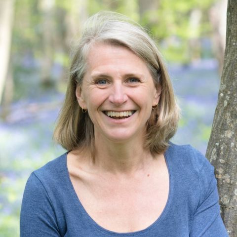 Jenevora Williams has grey-blonde hair in a long bob, blue eyes, and a kind smile. She's outside in a park and is wearing a scoop-neck duck egg blue top.