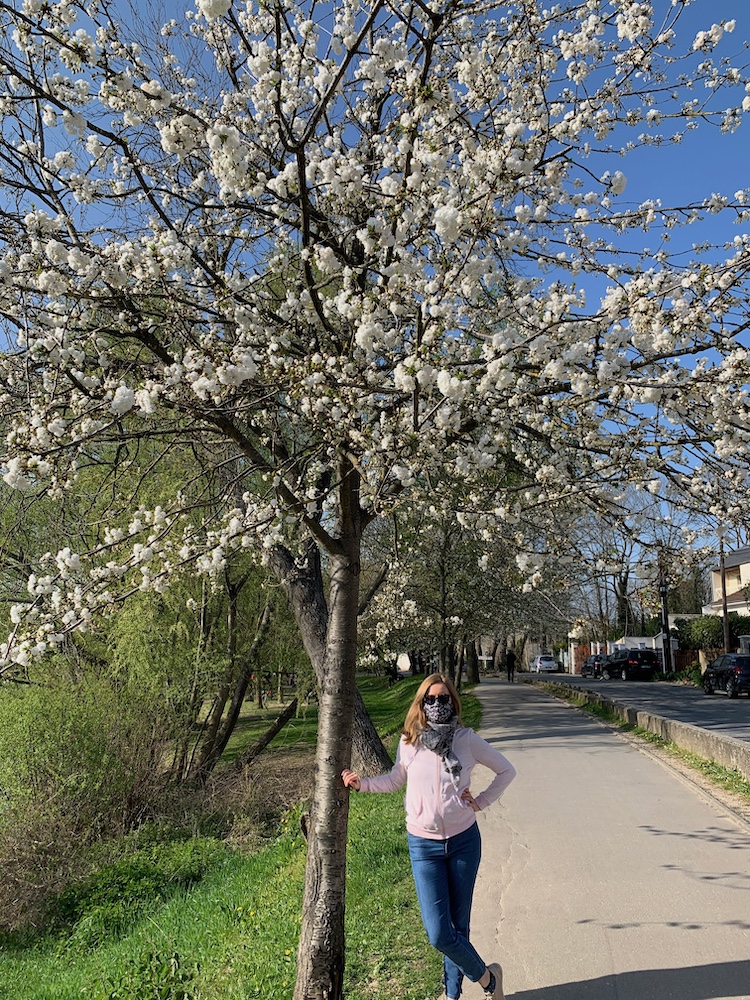 Georgia Aussenac standing under a white blossom tree next to a walkway, wearing a facemask and sun glasses to protect from seasonal allergies. It's a spring day: blue skies, green grass, but long sleeved pale pink hoody and blue jeans with sneakers.