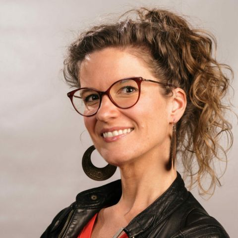 Sarah Algoet is a woman with curly chestnut hair, in a high ponytail. She's wearing strong framed red glasses, and big black hoop earrings, a black leather jacket and a red top. Her skin loves the sun, and she's got a mischievous smile.