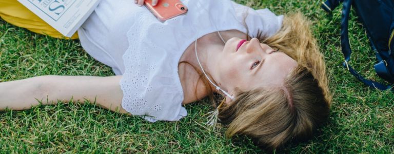 Georgia Aussenac is lying in the grass, listening to her phone through headphones, with a sheet music book across her belly. She's wearing a white lace top and a bright yellow skirt, and her blonde hair is fanned out over the grass. Her metallic navy blue backpack is to her right.