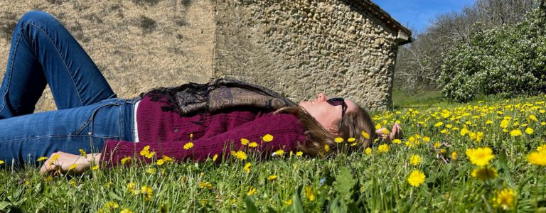 Georgia Aussenac is lying on a lawn surrounded by yellow daisies, in front of an old stone farm building. She's wearing a dark pink knit sweater, jeans and sunglasses, and she's relaxing looking up at the blue sky.