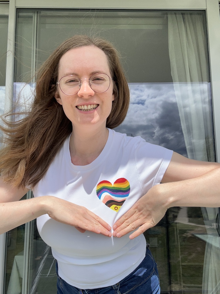Georgia is wearing a white tshirt with a painted heart with an updated pride flag rainbow. She's showing it off with her hands and smiling at the camera.