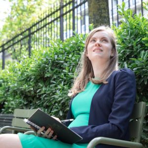 Georgia Aussenac, a white woman in a bright green short crepe dress and navy blazer, is sitting on a bench in a park writing in a notebook. She's looking up to the sky.