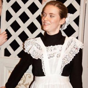 Georgia Aussenac, a white woman in her early 20s, is performing as Susanna in the Marriage of Figaro. She's leaning against a lattice fence as she sings, dressed in black and white as a maid.