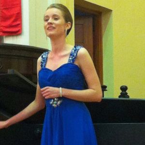 Georgia Aussenac, a white woman in her early 20s, is singing in a church in a formal electric blue bejewelled gown and her hair in a formal updo. She's singing, one hand in front of her waist.