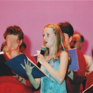 Georgia Aussenac is a white ten year old girl, singing with a group of adults on a stage with a pink backdrop, holding a mic. She looks confident.
