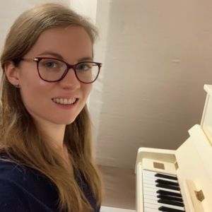 Georgia Aussenac, a white woman with long blonde hair and red framed glasses, is sitting at a white upright piano, turned towards the camera with a smile.