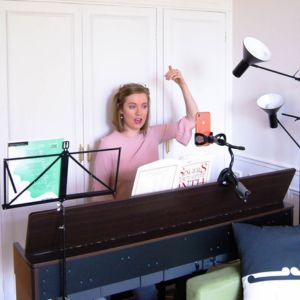 Georgia Aussenac, a white woman with short blonde hair, is sitting at a keyboard with a music stand to one side, and a phone on a tripod in front of her, singing a voice exercise with one hand raised about her head in gestural demonstration.